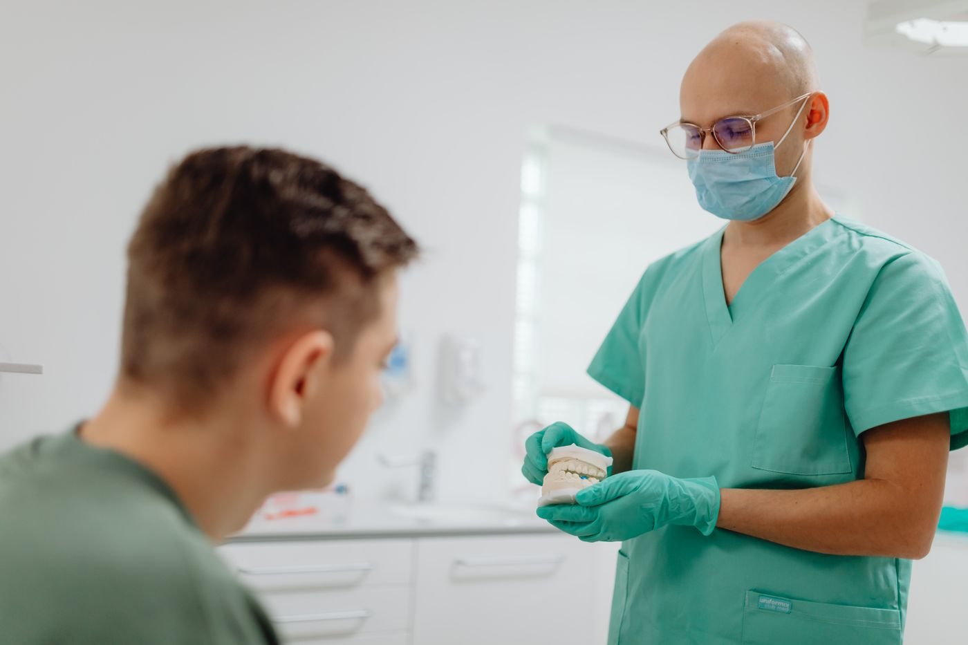 A dentist standing beside a seated patient, holding a model of the teeth and explaining what a treatment involves.